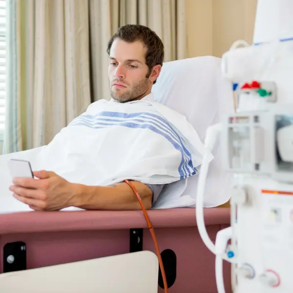 Patient reclining in a dialysis treatment chair while holding a smartphone, with medical equipment nearby