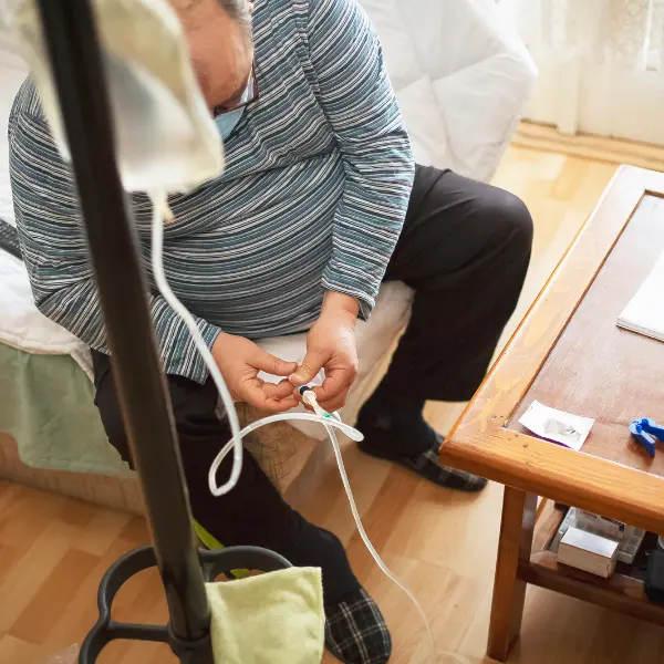 Person seated at home handling medical tubing beside a stand